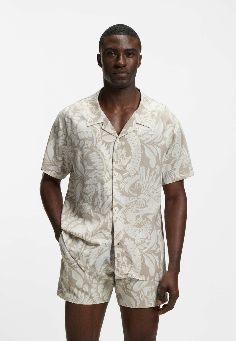 Homme portant une chemise à manches courtes boutonnée beige et blanche ainsi qu'un short assorti avec un imprimé de feuilles tropicales et de fleurs, debout devant un fond blanc.