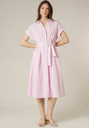 Femme debout, les mains sur les hanches, portant une robe midi ceinturée à rayures roses et blanches et des sandales à talons blocs dorées à bouts ouverts.