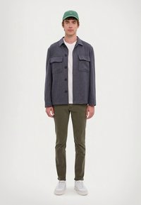 Šedá košile na knoflíky se dvěma náprsními kapsami přes bílé tričko, v kombinaci s olivově zelenými kalhotami a bílými teniskami, doplněno zelenou čepicí.