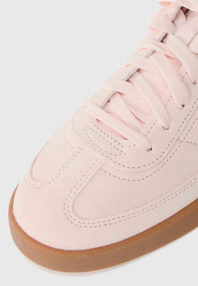 Zapatilla de ante rosa claro con una superficie lisa, costuras tonales y suela de goma. Presenta cordones planos y un diseño de puntera redondeada.