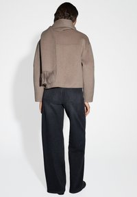 Beige wollen mix jas met een hoge kraag, gecombineerd met donkere wijde jeans. De jas heeft een gedrapeerde sjaal en strakke lijnen.