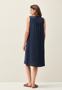 Mouwloze marineblauwe jurk met een zachte textuur, vloeiende silhouet en een geraffineerde achterkant, gecombineerd met bruine instapsandalen.