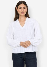 Hvid bluse med V-hals, tekstureret rynkning og bløde lange ærmer. Bæres med mørke jeans, der fremhæver en løs og afslappet pasform.