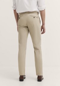 Beige Hose mit taillierter Passform, ausgestattet mit zwei Gesäßtaschen mit Knopfverschluss, aus einem glatten Stoff gefertigt. Getragen mit braunen Schuhen.