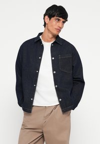 Temno modra denim jakna s gumbom spredaj in enim žepom na prsih, v kombinaciji z belo majico in bež hlačami. Gladka tekstura in strukturiran dizajn.