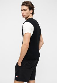 Μαύρο πλεκτό αμάνικο πουλόβερ με ριμπ υφή, σε συνδυασμό με μαύρα σορτς που έχουν ένα λογότυπο σε κόκκινο, άσπρο και μπλε στο πλάι.