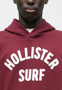 Bordo dukserica s bijelim izvezenim natpisom "HOLLISTER SURF", s mekanom teksturom i kapuljačom sa vezicama.