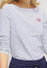 Randig långärmad topp i vit och marinblå, med en bred halsringning och röd "Polo" broderi på vänster sida. Mjuk bomullsmaterial.