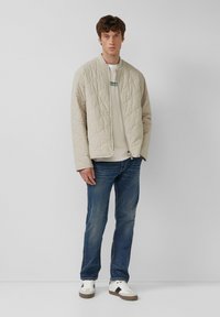 Beige quilted bomber jacket with a stand collar, paired with a light gray sweatshirt, blue jeans, and white sneakers with dark accents.