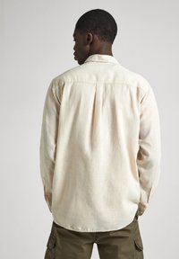 Beige long-sleeve shirt in lightweight fabric, featuring a buttoned collar, single back pleat, and cuffs, paired with olive cargo pants.