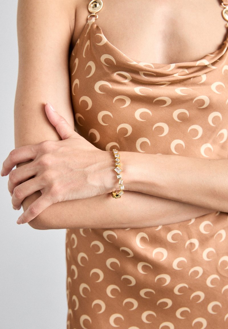 Χρυσό βραχιόλι με γεωμετρικές κρυστάλλινες λεπτομέρειες, που διαθέτει μίγμα σχημάτων. Το βραχιόλι φοριέται σε ένα ανοιχτόχρωμο χέρι, με φόντο υφασμάτινο με πατίνα.