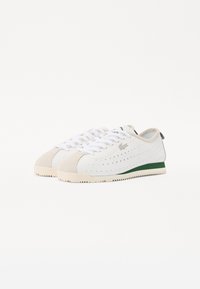 Valkoiset nahka-sneakerit, joissa on beige mokkanahka-korostuksia, vihreä kantapääyksitys, rei'itetyt sivut ja valkoiset nauhat. Teksturoitu kumipohja, joka tarjoaa hyvän pidon.