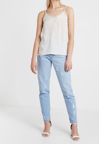 Bílý saténový top s krajkovým lemem, spárovaný se světle modrými džínami s vydřeným vzhledem. Džíny mají volný střih a třepený lem. Jednoduché sandály na tenkých páscích.