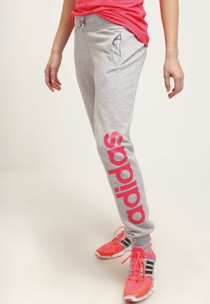 Osoba ubrana w szare dresy Adidas z różowym logo i różowe buty sportowe, stojąca na jednolitym jasnym tle.
