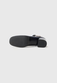 Scarpa nera in pelle con tacco, punta rotonda, tacco spesso e una texture liscia. Presenta un semplice fiocco decorativo sul lato.