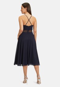 Tengerés kék A-vonalú ruha állítható, keresztbe tett pántokkal, csipke panellel a deréknál és laza szoknyával. Ezüst magassarkú szandálokkal viselve.