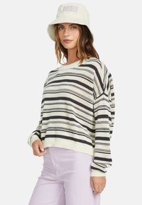 Striped sweater in cream and black features a loose fit, textured knit, and ribbed cuffs. Paired with light purple pants and a bucket hat.