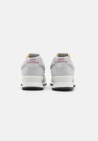 Harmaat mokkakengät valkoisella pohjalla. Ominaisuuksina keltainen ja harmaa kantapää sekä punainen "new balance" -logo takana. Minimalistinen muotoilu.