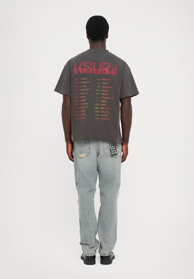 Graues Grafik-T-Shirt mit rotem Text auf dem Rücken, kombiniert mit lässigen hellblauen Jeans, die Patches und ein schwarzes Etikett an der Tasche haben.