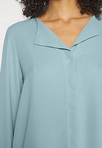 Světle modrá blůza s výstřihem do V, hladkou texturou a dlouhými rukávy. Má minimální prošití na přední straně.