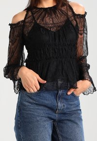 Blusa nera in pizzo con design off-shoulder, maniche allungate, vita arricciata e intricati motivi floreali. Abbinata a jeans blu.