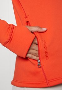 Kirkas oranssi, tiukasti istuva venyvää pulloveria, jossa on vetoketjullinen tasku ja brodeerattu logo. Ominaisuuksiin kuuluu sileä pinta ja peukalonrei'illä varustetut hihansuut.