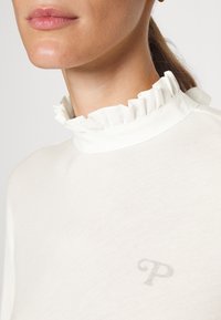 Vit långärmad topp med hög, ryschad krage och subtil "P"-logotyp framtill. Mjuk tyg med slät textur och åtsittande design.