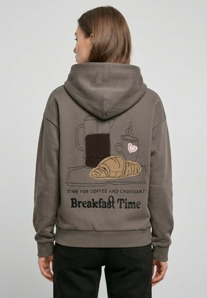 Frau trägt braunen Hoodie mit Rückillustration einer Kaffeekanne, dampfender Tasse mit Herz, Croissant und Text "Breakfast Time."
