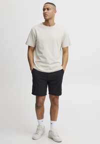 Beige bomull t-skjorte med korte ermer, kombinert med svarte shorts. Brukt med hvite sokker og joggesko. Merkbar tatovering på leggen.