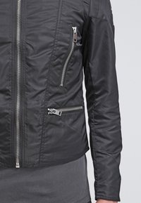 Svart zip-jacka med en lätt blänkande yta. Har flera zip-fickor och en figursydd design med kviltad textur på ärmarna.