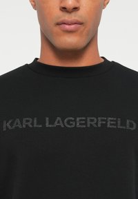 Črna bombažna majica s kapuco z rebrastim ovratnikom in embosiranim logotipom "KARL LAGERFELD" spredaj, ki se ponaša z gladko teksturo.