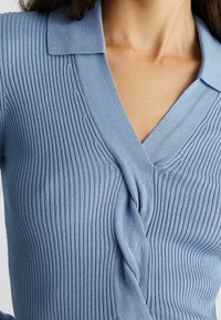 Ljusblå stickad tröja med ribbad struktur, V-ringning och krage. Har en snodd tygdetalj i mitten.