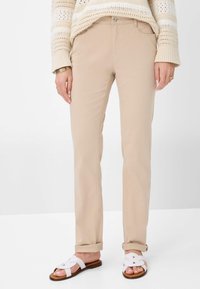 Beige, rechtvallende broek gemaakt van lichte stof, met een vlakke voorkant, riemlussen en opgerolde mouwen. Draagt witte sandalen.
