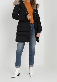 Musta toppatakki, jossa on turkiksella reunustettu huppu, vetoketjusuljenta ja tikattu muotoilu; puettu oranssin neuleen päälle, yhdistettyyn käännettyihin farkkuihin ja valkoisiin saappaisiin.