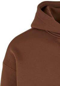 Brun hettegenser med et mykt, strukturert stoff, med en stor halsåpning og synlig søm. Designet er enkelt og moderne.