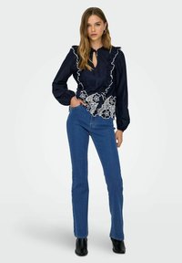 Blusa azul marino con detalles de encaje blanco en el dobladillo, escote volante, mangas largas y cierre con lazo. Combinada con jeans azul oscuro de cintura alta.