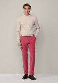Homme se tenant debout, portant un pull en tricot beige, un pantalon rouge et des bottes à lacets marron foncé devant un fond gris uni.