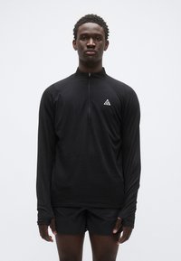 Top preto de manga comprida com fecho de correr, feito de tecido texturizado, com um pequeno logótipo branco no peito esquerdo, combinado com calções pretos.