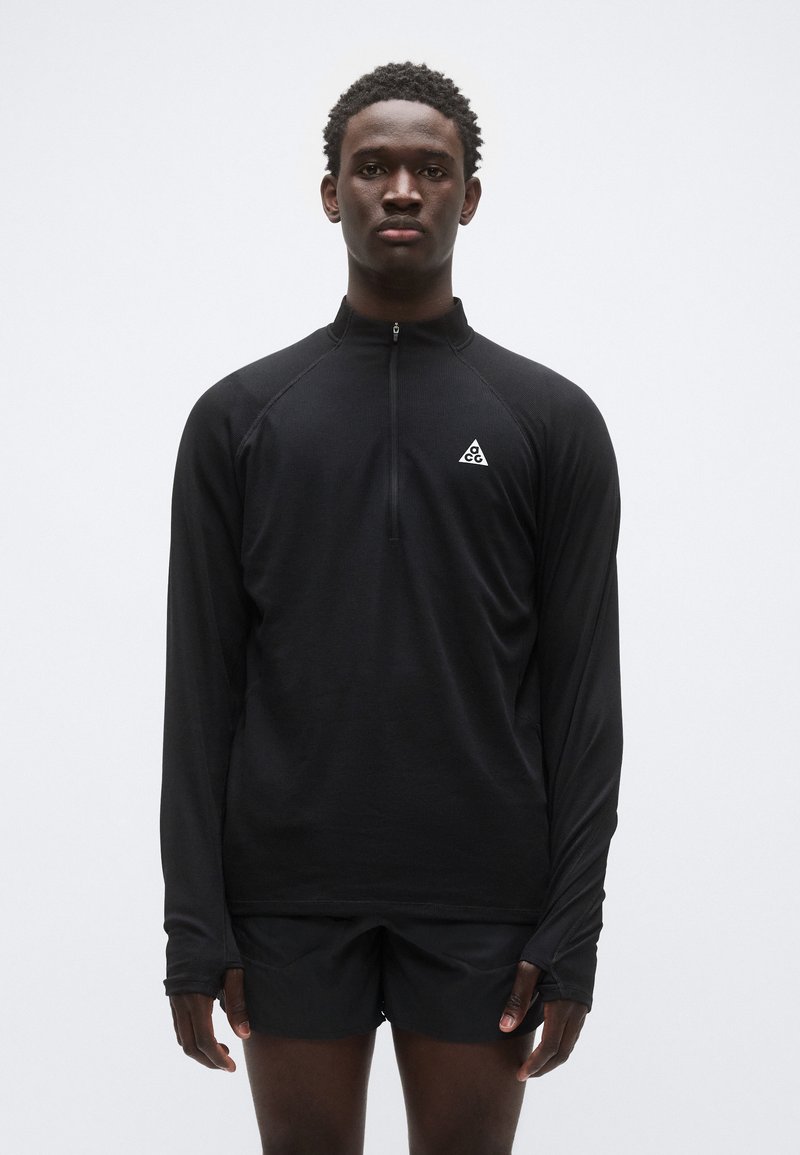 Top preto de manga comprida com fecho de correr, feito de tecido texturizado, com um pequeno logótipo branco no peito esquerdo, combinado com calções pretos.