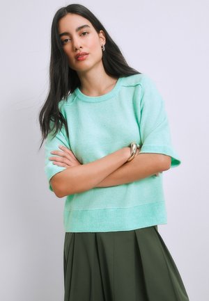 Vrouw met lang donker haar die een licht turquoise kort mouwen trui draagt en een donker olijfgroene rok, staand met gekruiste armen.