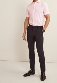 Homme debout portant une chemise à manches courtes rose clair à boutons, un pantalon slim foncé, des chaussures de ville noires et une montre-bracelet.