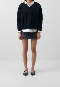 Navy cable knit sweater with a v-neck, white collared shirt underneath, grey denim mini skirt, and black shoes with studded accents.