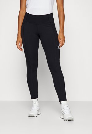 Frau trägt schwarze Adidas-Leggings mit hohem Bund, weiße Sportschuhe und ein weißes ärmelloses Top, steht vor einfachem Hintergrund.