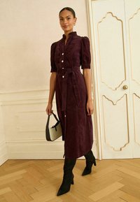 Burgundi värvi midi kleit, millel on volangkrae, puffilavad ja sidumisvöö, esifunktsiooniga nööbid. Mudel hoiab väikest musta ja valget kotti.