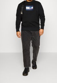 Černá mikina s modro-červenou grafickou nášivkou s nápisem "TOMMY JEANS." Kombinováno s šedými džínami a černými kotníkovými botami. Jednoduchý design a střih.