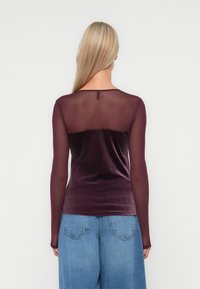 Langemouwtop met een diep bordeaux velours lijfje en doorschijnende mesh mouwen, met een ronde halslijn en een knoopdetail op de rug.