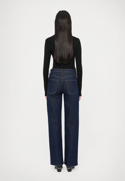 Tumesinised kõrge vöökohaga teksad, lõdva lõikega, kahel tagataskul ja nähtava õmblusega. Kandmisel on keha järgiva musta pikavarrukaga pluusiga.