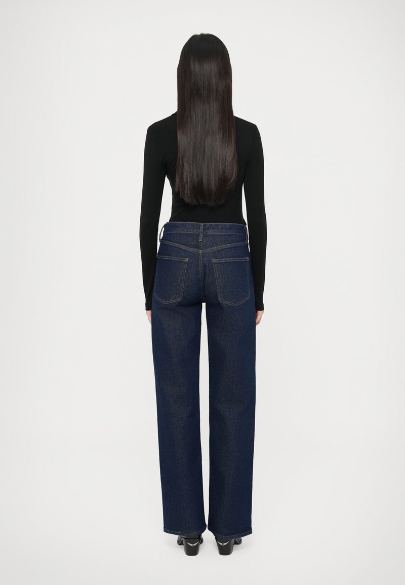 Dunkelblaue Jeans mit hoher Taille und lockerem Sitz, mit zwei Gesäßtaschen und sichtbaren Nähten. Kombiniert mit einem enganliegenden schwarzen Langarmoberteil.