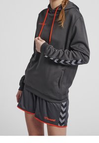 Grauer Hoodie mit orangefarbenen Akzenten und Logo, aus strukturiertem Stoff mit einer Kängurutasche. Kombiniert mit passenden Shorts im Chevron-Muster.