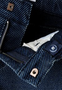 Tela de mezclilla azul oscuro con un zipper plateado visible y un botón de cobre. Presenta un complicado bordado y un forro blanco en el interior del bolsillo.